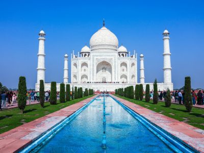 The iconic white marble Taj Mahal perfectly reflected in the garden's water channel during the soft light of sunrise, with clear blue skies in the background